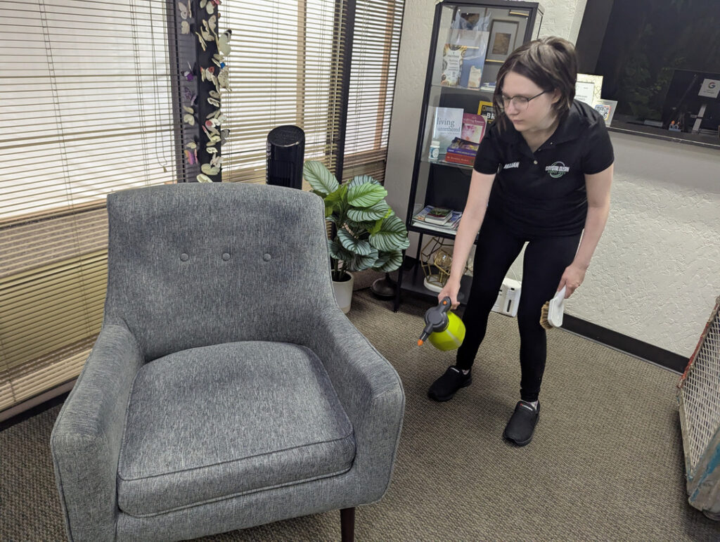 a woman cleaning a gray upholstered chair using a spray bottle and a brush in an office setting