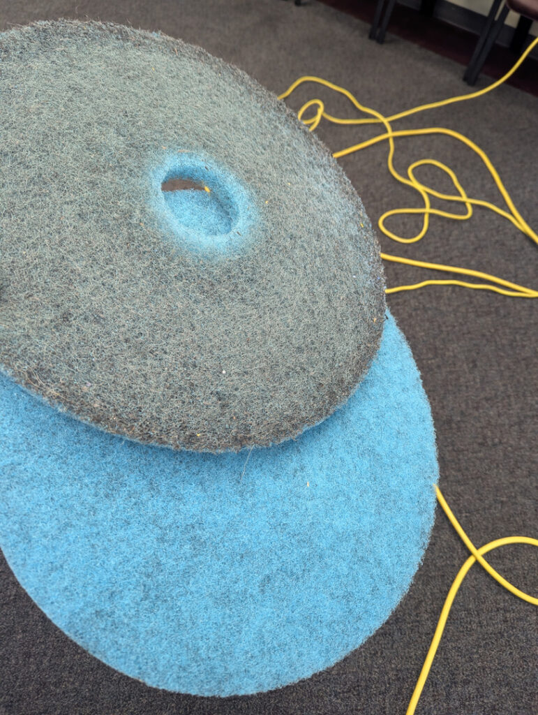 blue and gray carpet cleaning pads stacked on a dark carpet with a yellow electrical cord in the background