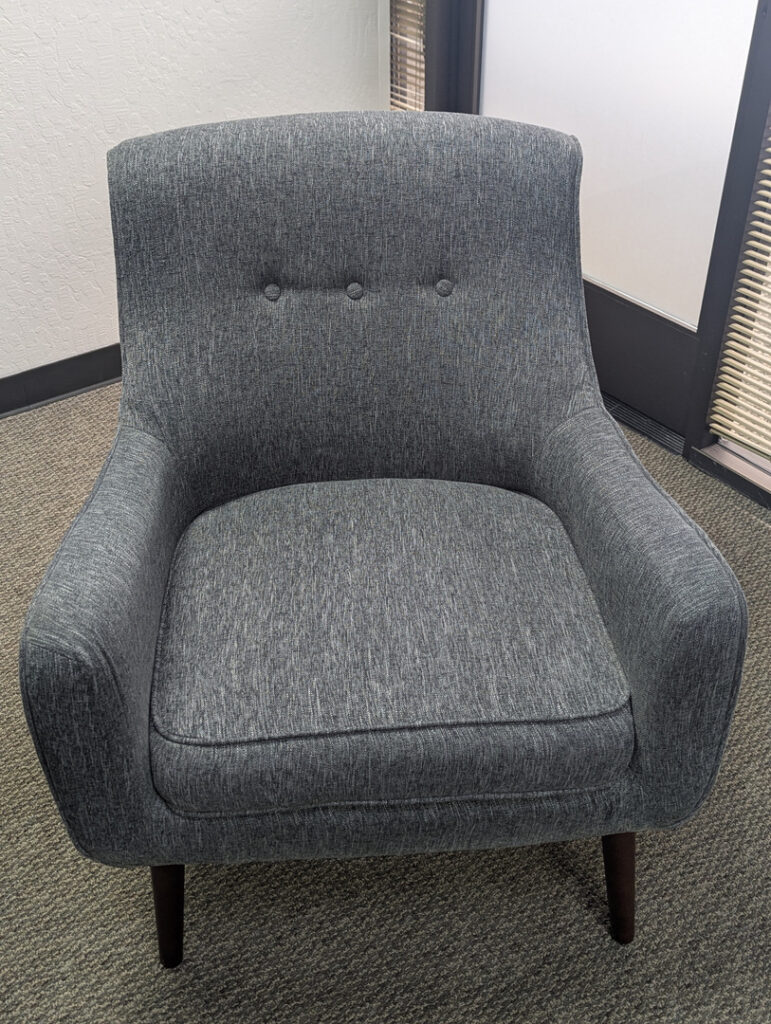 gray upholstered chair placed on a carpeted floor in a well-lit indoor setting with natural light coming through the window